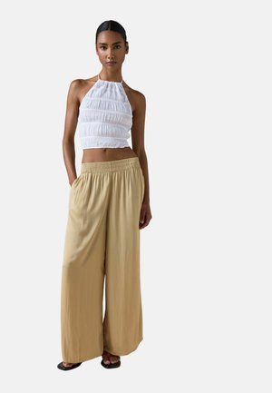 Kvinde iført hvid rynket haltercrop top og løstsiddende beige viddebenbukser står med hænderne i lommerne.