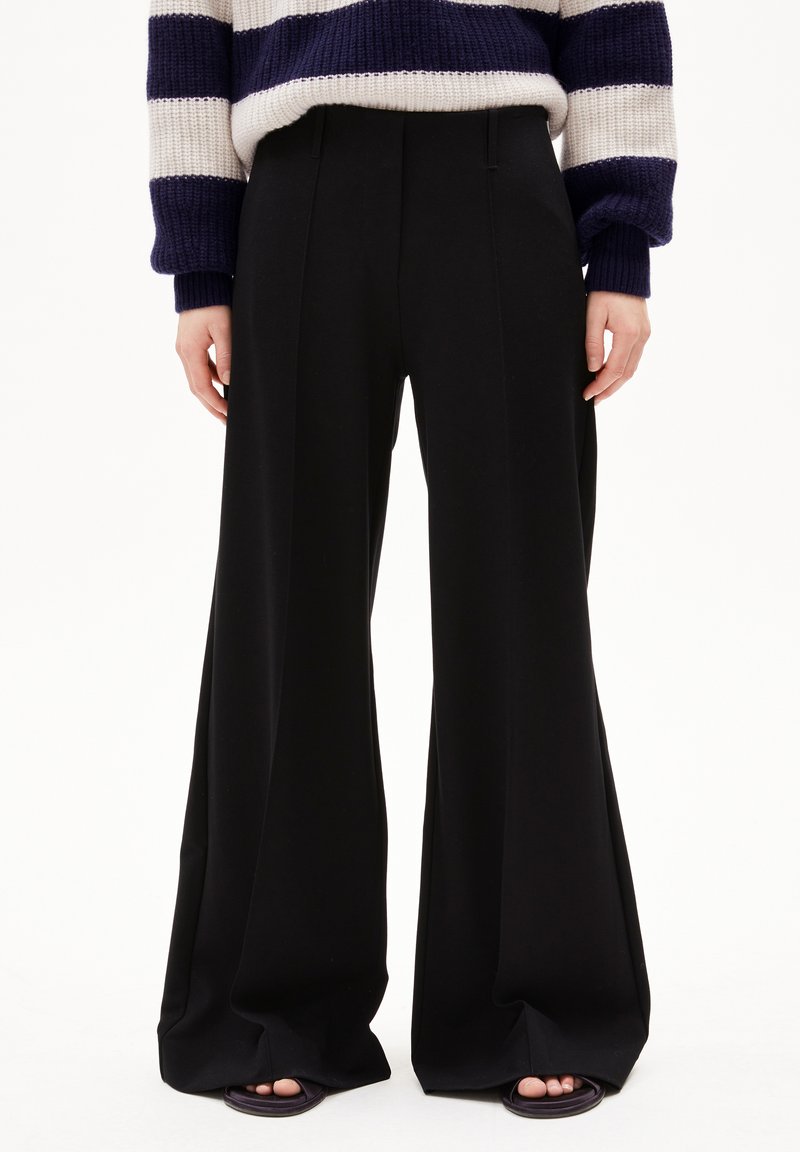 Wide-leg black trousers made from a smooth fabric with a clean design. They feature pressed creases and are worn with open-toed shoes.
