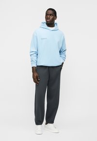 Muž stojící s rukama v kapsách, oblečený v světlomodré mikině, tmavě šedých teplácích a bílých teniskách, na jednobarevném pozadí.