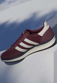 Baskets Adidas couleur marron avec des bandes blanches et un onglet au talon, positionnées sur une surface blanche devant un arrière-plan gris-bleu flou.