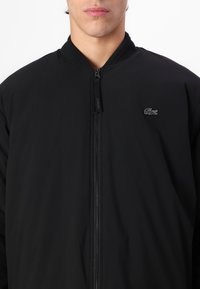 Jachetă bomber neagră cu guler ribbed și închidere cu fermoar. Prezintă un logo brodat subtil pe piept și o textură de țesătură netedă.