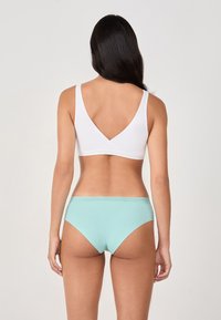 Frau von hinten gesehen, die ein weißes ärmelloses Crop-Top und hellblaue Bikiniunterwäsche trägt, vor einem schlichten Hintergrund.