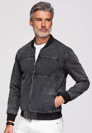 Mand med gråt hår og skæg iført en sort denim bomberjakke og hvide bukser, hånden i lommen, kigger til siden.