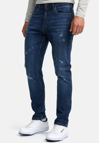 Jeans de mezclilla azul oscuro con un corte ajustado, que presentan detalles desgastados y acentos deshilachados. Combinados con zapatillas deportivas blancas.