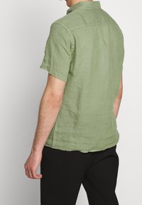 Lyhythihainen paita vaaleanvihreästä pellavasta, jossa on kauluskukkanen ja rento leikkaus. Rakenteellinen kangas, jossa on hienovaraisia ompeluyksityiskohtia.