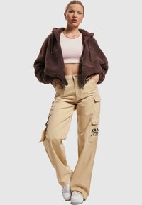 Brun shearling jakke med glidelås, lys rosa crop-top, beige cargo bukser med sidelommer, og hvite joggesko, som viser en avslappet passform og uformell stil.