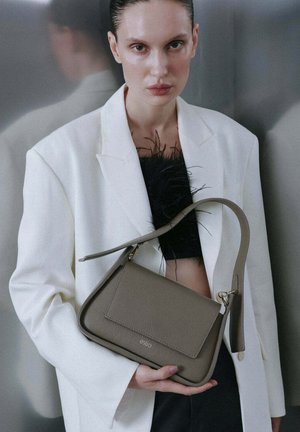 Femeie purtând sacou alb și top negru cu pene ținând o geantă de mână din piele taupe cu bretea de umăr pe fundal gri.