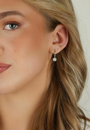 Silvriga hoops örhängen med en blommig snöflingedesign, prydda med klara ädelstenar, hängande under loben i en närbild.