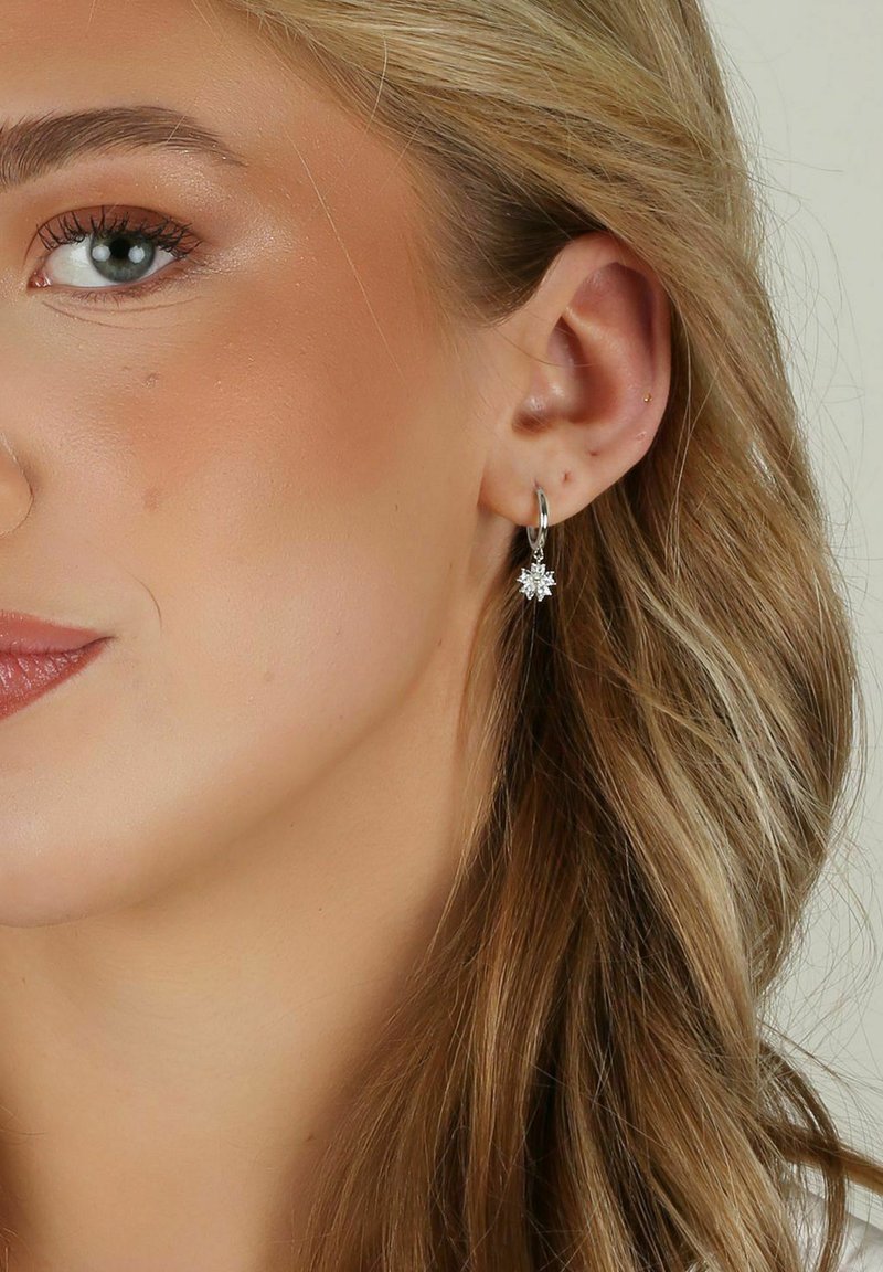 Silverörhängen i form av hoops med ett blomster- och stjärnliknande hänge dekorerat med små klara stenar, som hänger nedanför örsnibben. Slät textur, glänsande yta.