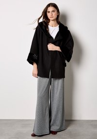 Schwarze Außenbekleidung mit Kapuze, die glänzenden Stoff an den Ärmeln besitzt. Kombiniert mit weit geschnittenen Hosen im Dogtooth-Muster und burgunderfarbenen Schuhen.