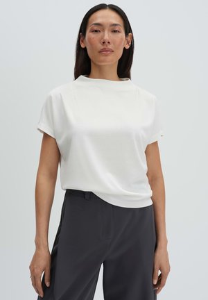 Donna con lunghi capelli scuri che indossa una maglietta bianca a maniche corte e pantaloni scuri, in piedi davanti a uno sfondo chiaro e uniforme.