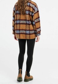 Chemise en flanelle à carreaux bruns avec des accents bleus et rouille ; coupe décontractée, manches longues et épaules tombantes. Associée à des leggings noirs et des bottes marron.