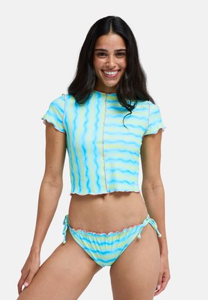 Junge Frau mit langen dunklen Haaren, lächelnd, trägt ein blau-gelb gestreiftes kurzes Top mit kurzen Ärmeln und passende Bikinihosen.