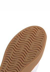 Suola di scarpa in gomma color sabbia con un motivo a onde, caratterizzata da una presa testurizzata. I bordi si curvano dolcemente verso un materiale superiore bianco.