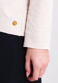 Ljust beige stickad tröja med ribbad manschett och en gyllene rund knappdetalj nära fållen, med en mjuk textur och avslappnad passform.