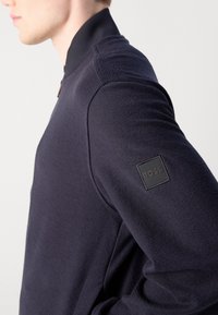 Vue latérale d'une personne portant une veste texturée bleu marine foncé avec un col montant et un écusson noir "BOSS" sur la partie supérieure de la manche.