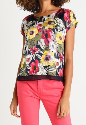 Woman wearing a short-sleeve floral top with red, yellow, and white flowers, paired with bright pink pants against a plain background.