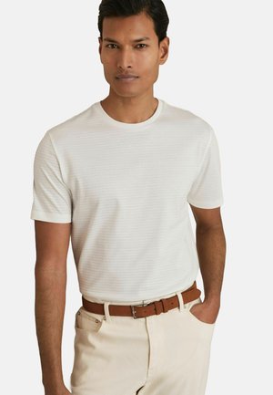 Camiseta blanca de manga corta con sutiles rayas verticales, cuello redondo, combinada con pantalones beige y un cinturón marrón. Textura de tela suave.