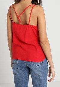 Top rosso senza maniche con spalline sottili e vestibilità morbida, realizzato in tessuto liscio, con orlo arrotondato e dettaglio della spallina incrociata sul retro.