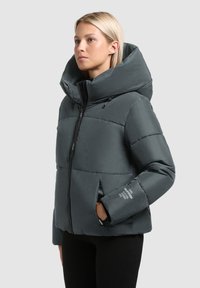 Kvinde iført en mørkegrå polstret jakke med høj krave og lynlås, står foran en ensfarvet lys grå baggrund.