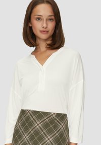 Weißes Longsleeve-Shirt mit V-Ausschnitt und Knopfdetails, kombiniert mit einem grün karierten Rock. Glatte Textur und lässige Passform.