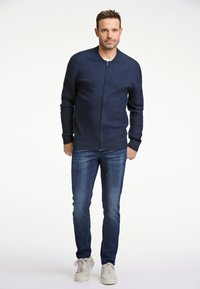 Marinblå zip-up cardigan med ribbade ärmslut och nederkant, i kombination med mörka jeans och ljusa sneakers, med en strömlinjeformad och figurnära design.