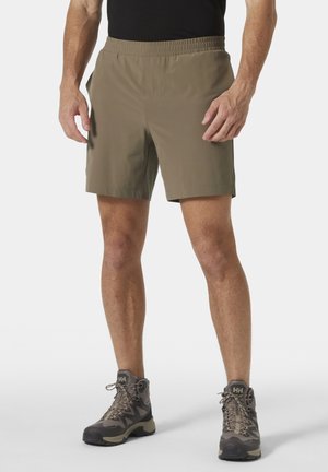 Man draagt olijfgroene sportshort en grijze wandelschoenen, staat met handen ontspannen langs het lichaam tegen een effen achtergrond.