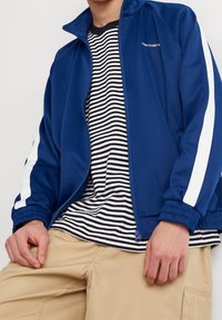 Henkilö, jolla on päällään sininen vetoketjullinen takki, jossa on valkoiset hihastripit, mustavalkoinen raidallinen paita ja beige cargo-housut, istuu kädet jaloillaan.