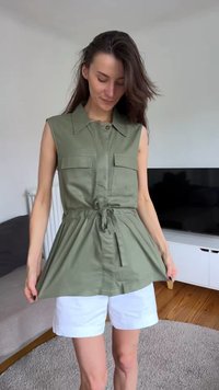 Femme portant un haut sans manches vert olive à boutons avec de grandes poches et un short blanc, debout dans un salon moderne.