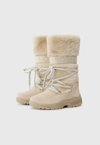 Beige szőrme bélésű csizmák vastag talppal és fűzővel, a tetején logószalaggal. A háttér egyszerű fehér, ami kiemeli a csizmákat.