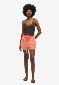 Brown knitted tank top with thin straps paired with high-waisted coral shorts featuring a tied belt and front pleats. Brown sandals.