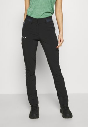 Femme portant un pantalon de randonnée noir avec des poches zippées, un t-shirt vert à manches courtes et des chaussures de randonnée noires, debout sur un fond blanc.
