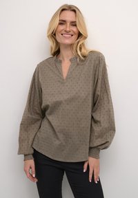 Olivově zelená blůza s volným střihem, texturovaným puntíkovaným vzorem a výstřihem do V. Má dlouhé rukávy se staženými manžetami.