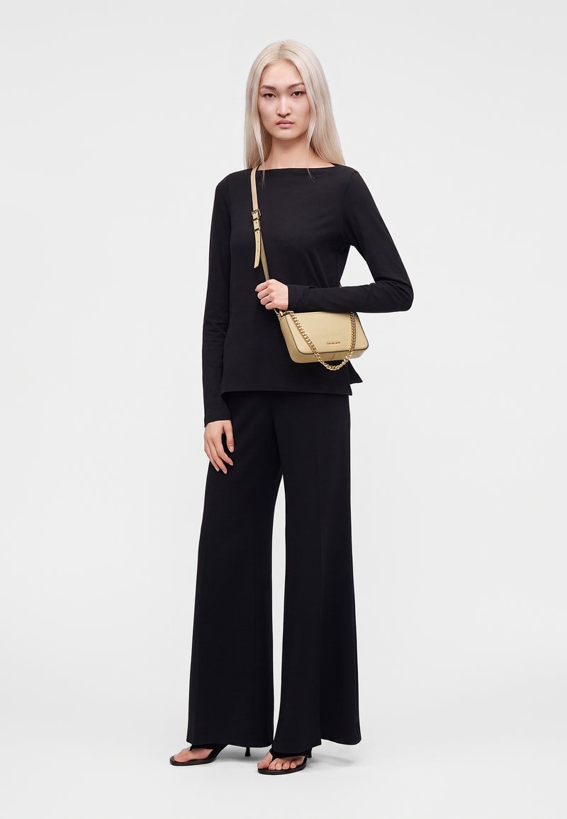 Jeune femme aux longs cheveux blonds, portant un haut noir à manches longues et un pantalon large, tenant un sac à bandoulière beige avec une chaîne dorée.