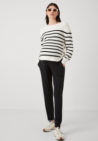 Hvit og svartstripete genser med knappeaccent, kombinert med svarte bukser og beige sneakers. Solbriller fullfører antrekket.