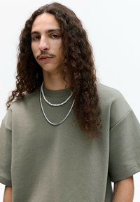 Olivengrøn oversized sweatshirt med korte ærmer, lavet af et struktureret stof. To lagdelte halskæder – en sølvfarvet og en perlekæde – pryder halsudskæringen.