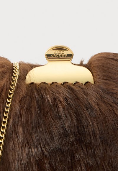 Καφέ τσάντα χειρός από ψεύτικη γούνα με μεταλλικό κούμπωμα σε χρυσό τόνο που φέρει το όνομα της μάρκας "Marc Jacobs." Το λουρί αλυσίδας προσθέτει μια αντίθετη υφή.