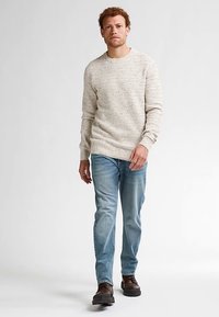 Een beige gebreide trui met een textuurpatroon, ronde hals en geribbelde manchetten, gecombineerd met lichtblauwe spijkerbroek en bruine schoenen.