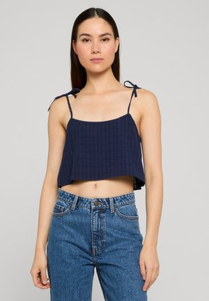 Junge Frau trägt ein marineblaues, gekürztes Tanktop mit gebundenen Schulterriemen und hoch taillierte blaue Jeans vor einem schlichten Hintergrund.