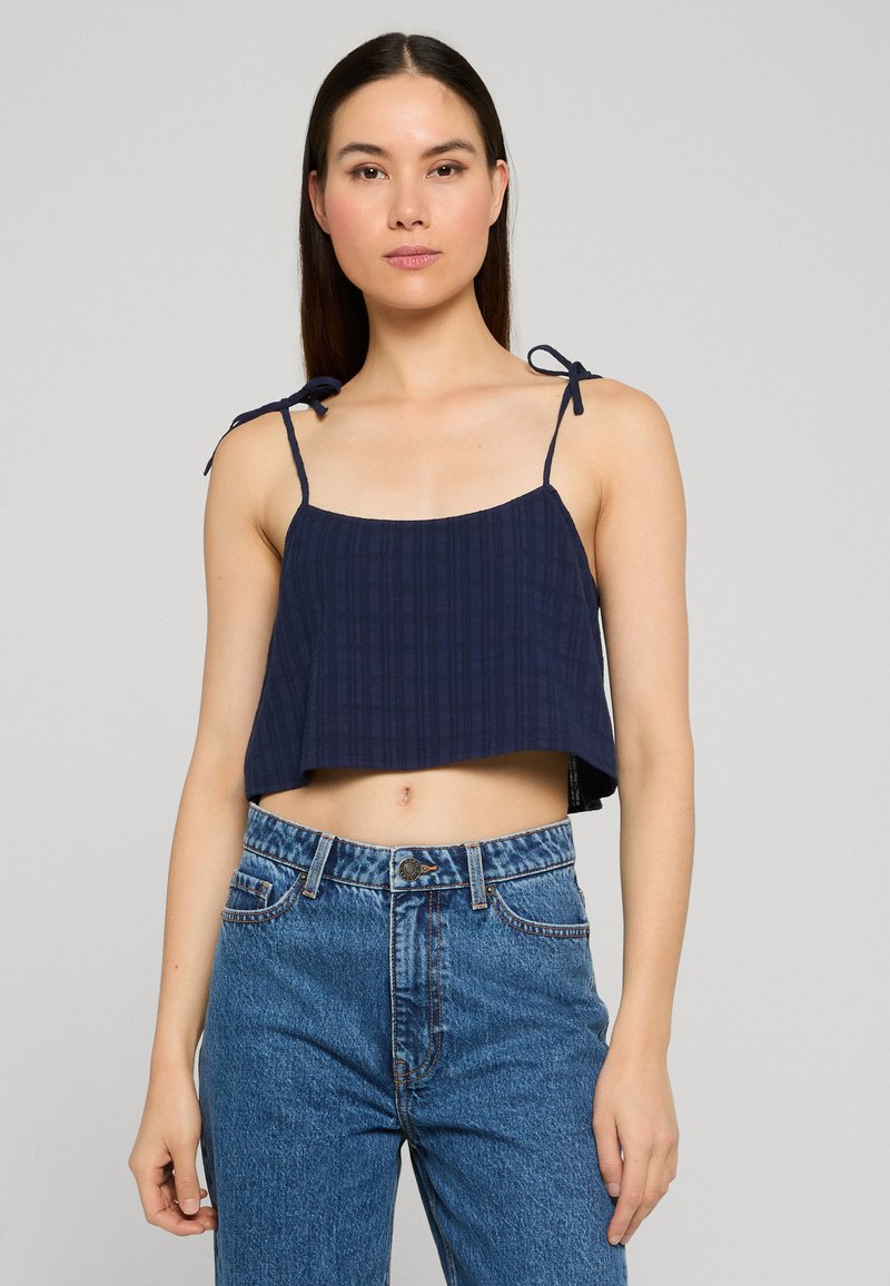 Jeune femme portant un débardeur court bleu marine avec des bretelles nouées sur les épaules et un jean bleu taille haute, sur un fond uni.