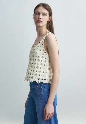 Ung kvinde iført ærmeløs cremefarvet hæklet top med blomstermønster og blå jeans, stående mod en ensartet lys baggrund.