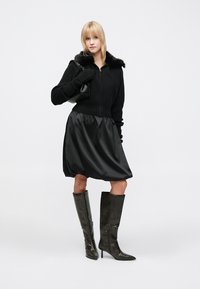 Jachetă neagră tricotată cu guler de blană, asortată cu o fustă neagră din satin. Modelul poartă cizme din piele gri închis până la genunchi. Geantă neagră plată inclusă.