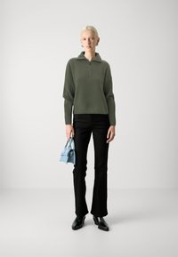 Olivgrüner gerippter Pullover mit Kragen und halbem Reißverschluss; kombiniert mit schwarzen weit geschnittenen Hosen und einer kleinen hellblauen Handtasche. Schwarze Stiefeletten mit Absatz.