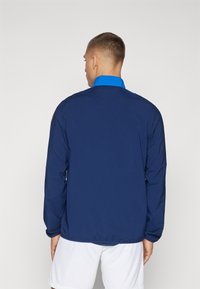 Giacca a vento blu con maniche lunghe, colletto alto e orlo elastico. Presenta un colletto a contrasto e un design minimalista.