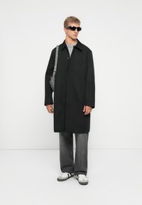 Joven con abrigo largo negro, camisa gris, pantalones anchos grises, zapatillas blancas, gafas de sol negras y llevando un bolso de hombro gris, de pie.