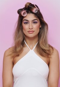 Modelo con cabello largo y ondulado, usando rulos de cabello rosa; tiene sombra de ojos púrpura sobre un fondo neutro y viste una blusa sin mangas blanca.