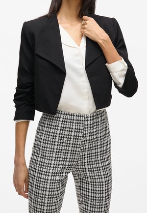 Vrouw draagt een korte zwarte blazer, een witte blouse met V-hals en een hooggetailleerde zwart-witte pantalon met pied-de-poule patroon.