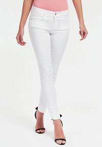 Jeans blancos de corte ajustado con cinco bolsillos, tejido ligero, diseño minimalista y un pequeño botón de metal en la cintura. Combinados con sandalias negras.