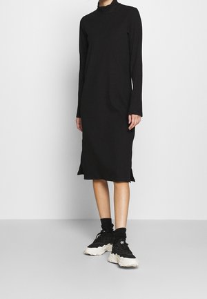 Kobieta ubrana w czarną, prążkowaną sukienkę midi z długim rękawem i rozcięciami po bokach, zestawioną z czarno-białymi masywnymi sneakersami i czarnymi skarpetkami.