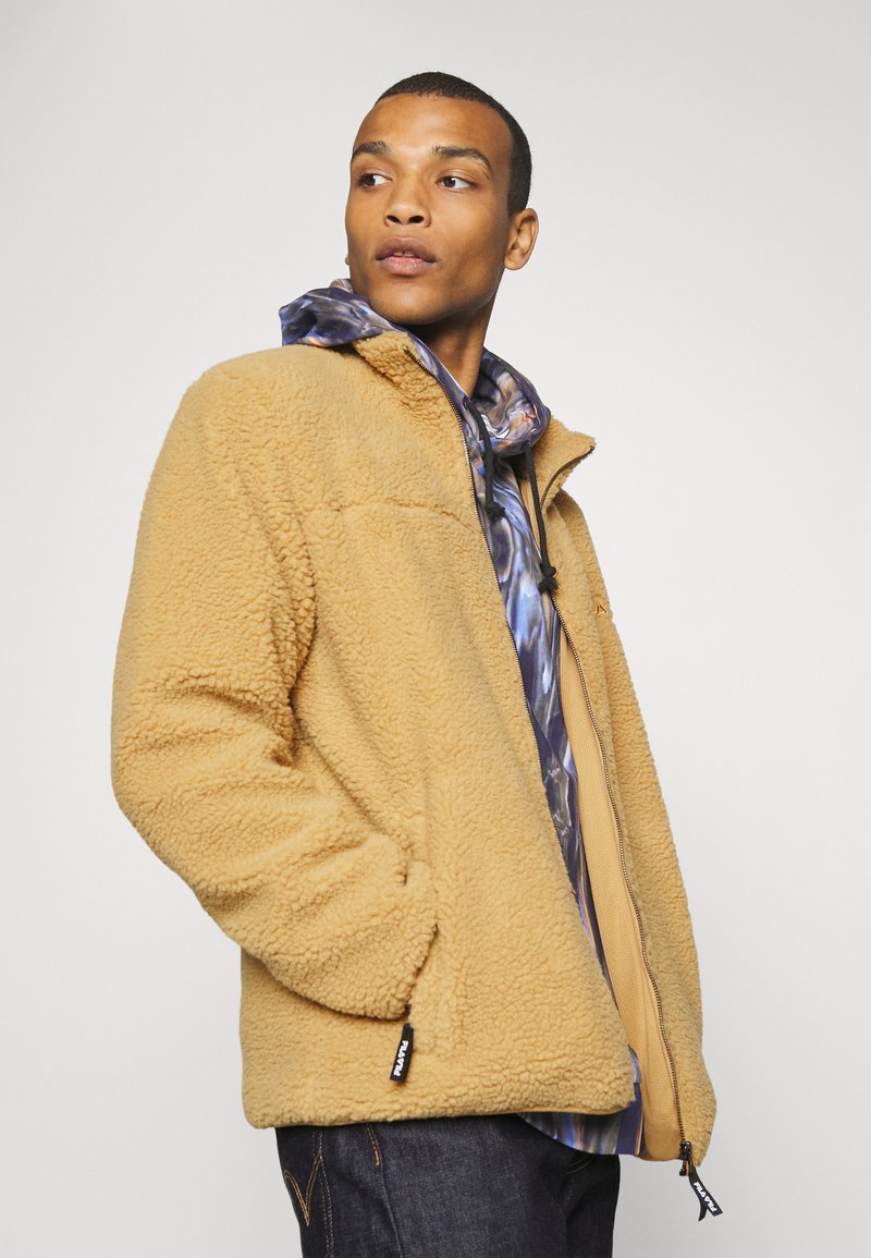 Giacca in pile beige con zip frontale, tasche laterali e una texture morbida, indossata sopra una camicia a cappuccio multicolore e decorata.